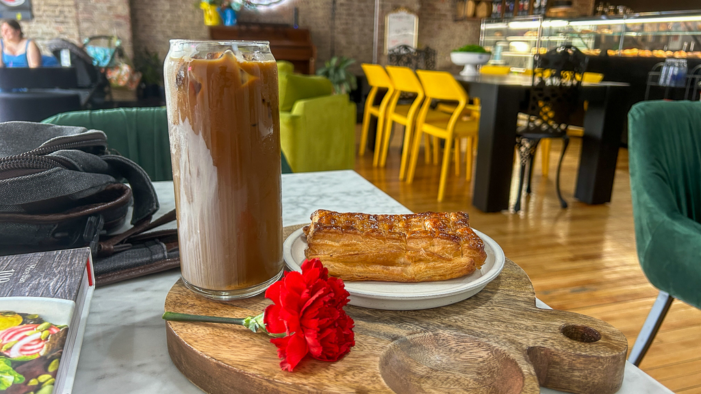 Dulce café corner with a tall coffee and a flaky pastry on a wooden board, accented with a red flower, against a backdrop of vibrant green chairs and black tables.